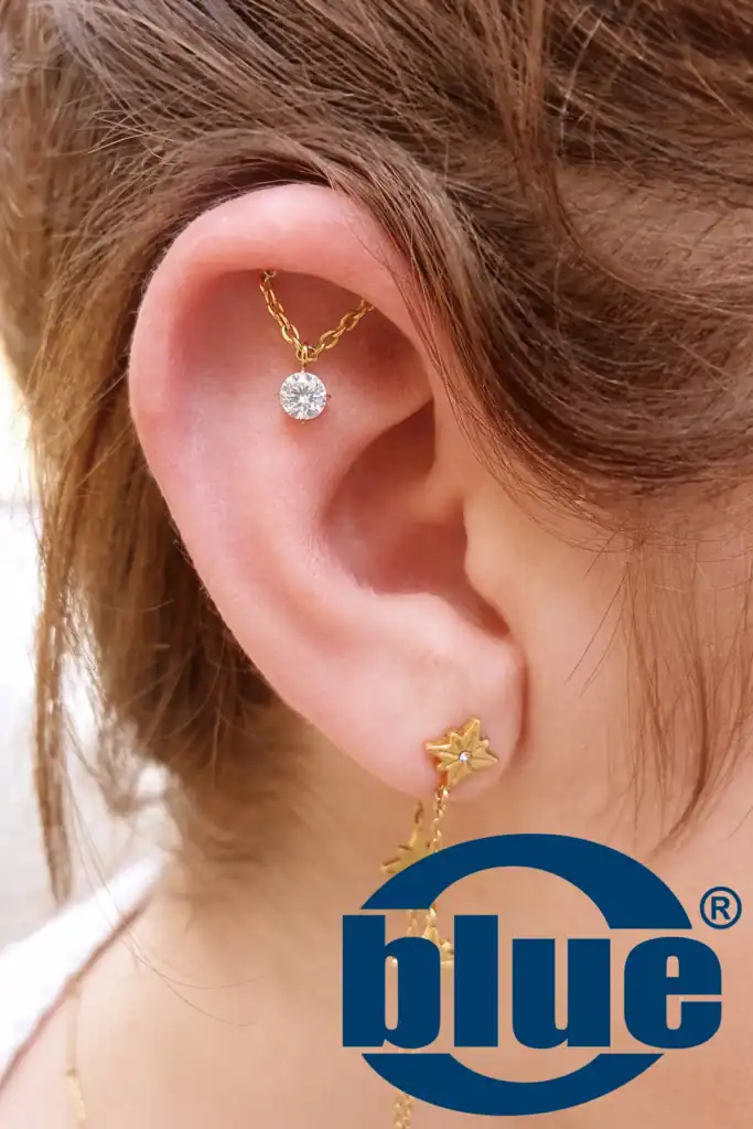 Close-up of a woman's ear with a gold chain and a small diamond stud on the upper cartilage; a gold star earring is on the earlobe, with a blue logo overlay in the lower right.