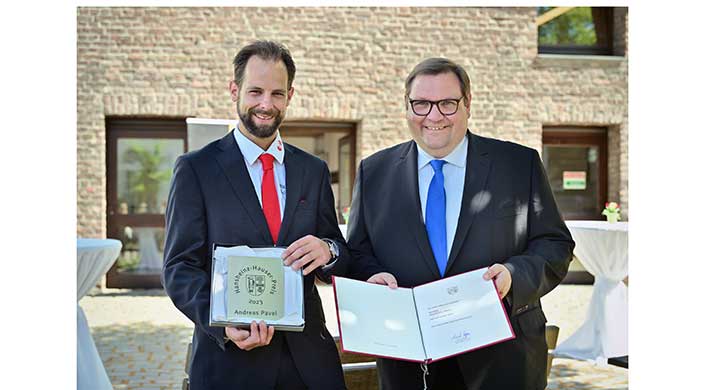 Zwei Männer im Anzug stehen draußen und halten aufgeschlagene Mappen in den Händen, beide lächeln in die Kamera.