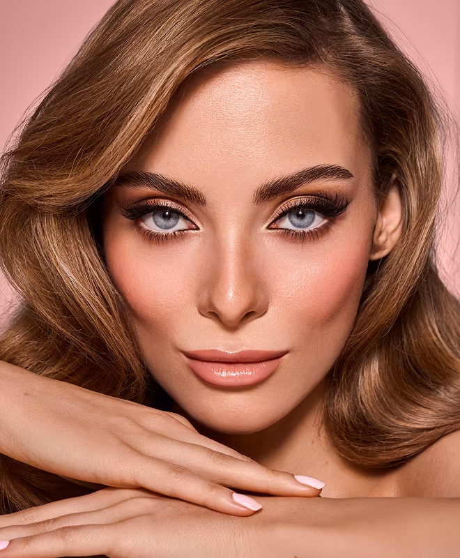 Frau mit Make-up und betonten Augen, weicher Haut und voluminösen Haaren auf rosa Hintergrund.