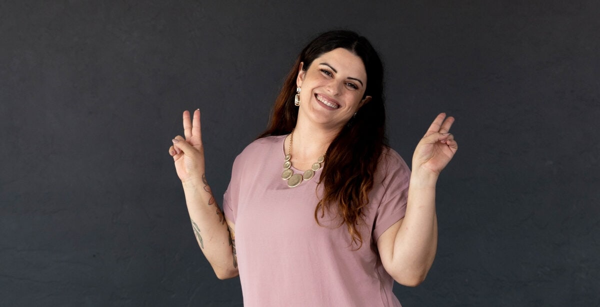 A smiling woman in a pink blouse and white pants poses with peace signs in front of a dark gray wall, featured on Canvas Rebel.