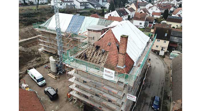 Mehrere Arbeiter renovieren ein Dach auf einem eingerüsteten Gebäude, teils mit entfernten Ziegeln. Im Hintergrund sind weitere Häuser zu sehen.