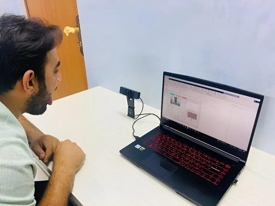 A man uses an AI tongue scan to analyze his health. He sticks his tongue out toward a camera connected to a laptop, which displays an AI tongue analysis interface. The image showcases a non-invasive diagnostic tool for potential disease diagnosis.