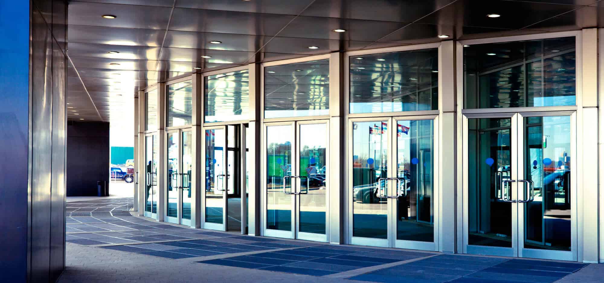 The image shows a modern building entrance with a row of glass doors, some of which are revolving doors. The entrance has a sleek design with metallic frames and a covered pathway leading to the doors. A locksmith can be seen working near the entrance, while people and vehicles are visible in the background.