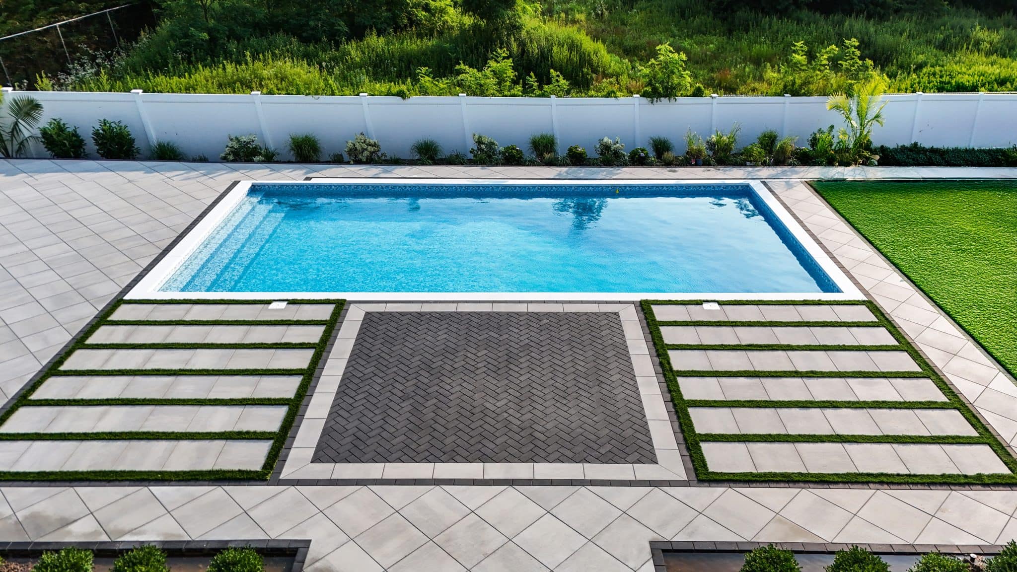 A rectangular swimming pool in a modern backyard, surrounded by paved tiles, geometric grass patterns, and a white privacy fence.