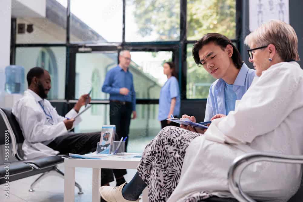 Asian patient holding clipboard signing medical insurance while elderly medic explaining health care treatment for curing ill. Multi ethnic people standing in hospital waiting area during consultation