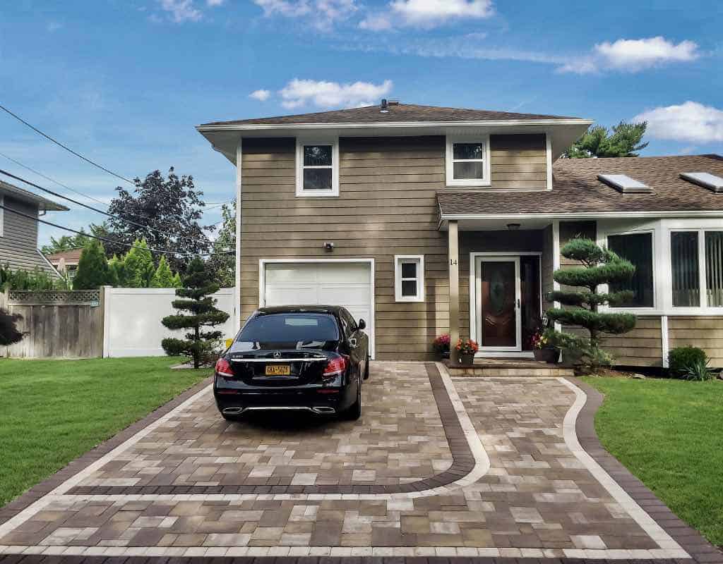 Two-story house with a brown facade, white trim, and a single garage. A black car is parked on the paved driveway. Neatly trimmed shrubs flank the entrance. Sunny day with a blue sky.