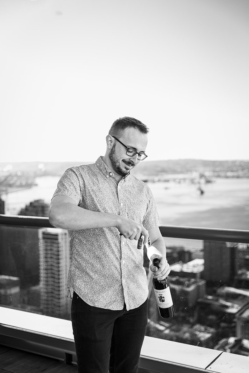 A man in glasses is opening a bottle on a balcony with a city and river view in the background.