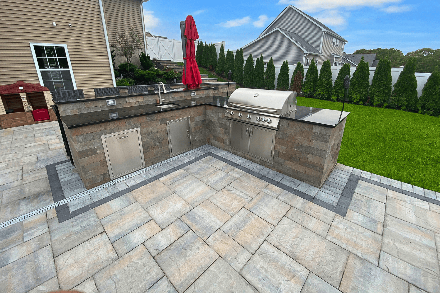 Outdoor Kitchens shine with a stainless steel grill, sink, storage cabinets, and a red umbrella on a stone patio surrounded by grass and privacy trees in the backyard.
