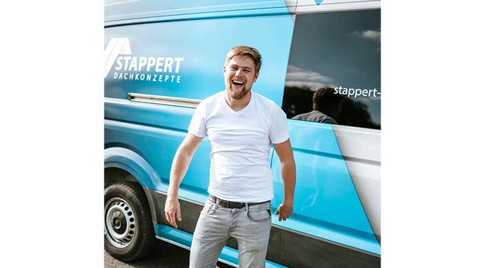 Ein junger Mann mit Bart und weißem T-Shirt lacht und steht vor einem blauen Lieferwagen.