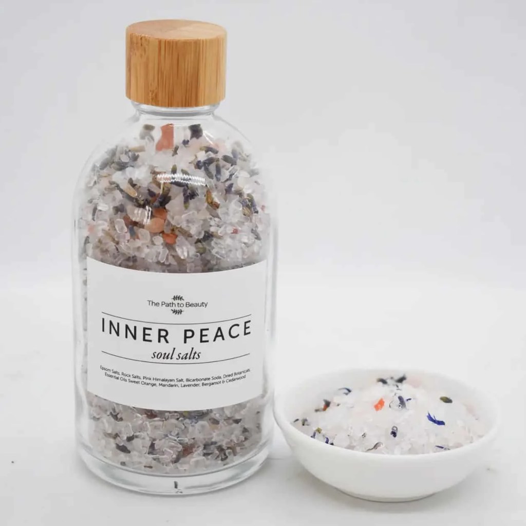 A clear glass bottle labeled Inner Peace Bath Salts with a wooden lid is placed beside a small white bowl filled with these colorful bath salts, which feature mixed minerals and dried flower petals.