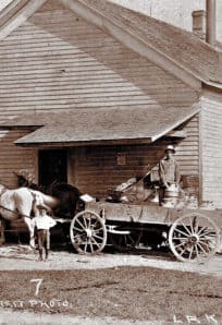 Creamery in Lakeville - Casad Horse-drawn milk delivery cart outside a wooden building in 19th-century rural America.