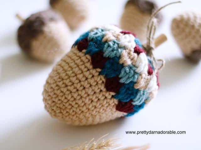 Close-up of a beige-based crocheted acorn with a blue, burgundy, and cream cap among other acorns; www.prettydarnadorable.com appears. Free crochet pattern