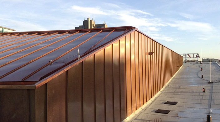 Metallisches Dach mit senkrechten Paneelen im Sonnenlicht, Gebäude und blauer Himmel im Hintergrund.