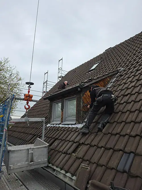 Zwei Dachdecker reparieren das Dach eines Hauses mit braunen Dachziegeln, ein Kranausleger und ein Gerüst sind ebenfalls zu sehen.