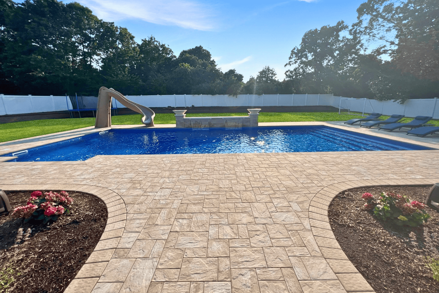 A rectangular outdoor swimming pool with a slide, surrounded by stone patios, lounge chairs, and landscaped garden beds, set against a backdrop of trees and a white fence.