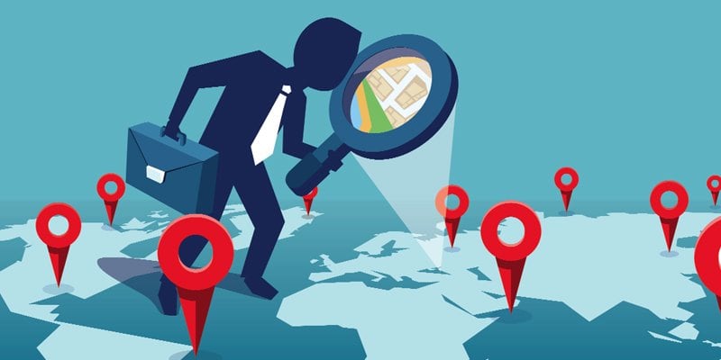 An illustrated graphic of a businessperson with a briefcase, holding a large magnifying glass over a world map. The magnifying glass reveals a zoomed-in view of a map with a specific location highlighted. Red location pins are scattered across the map, symbolizing various points of interest. The image conveys the concept of global navigation, location search, or business expansion.