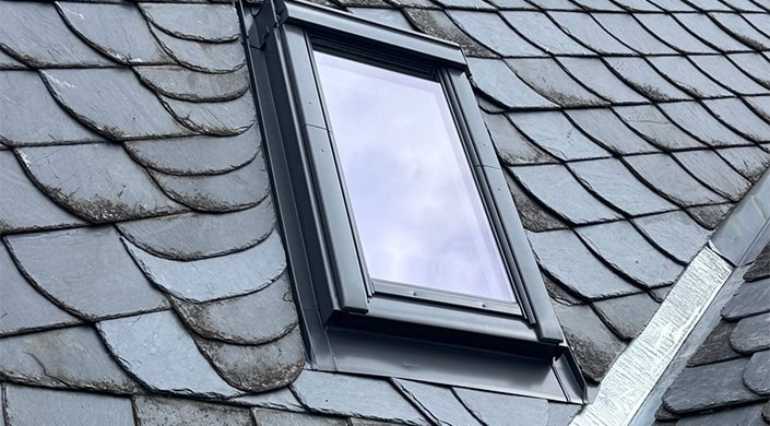 Schräges Dachfenster mit dunklem Rahmen, das den Himmel reflektiert. Das Fenster ist von grauen Dachziegeln umgeben.