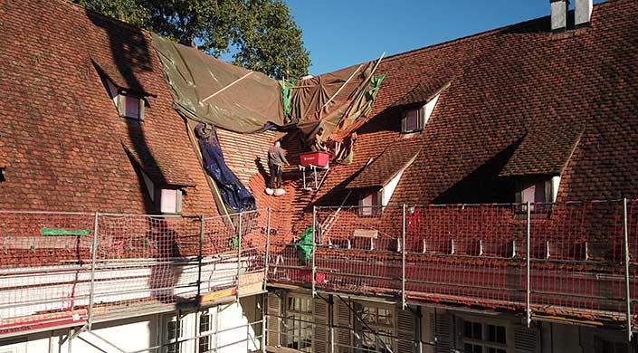 Zwei Bauarbeiter arbeiten mit einer Schubkarre auf einem Gerüst an einem schrägen Hausdach, das mit Planen abgedeckt ist.