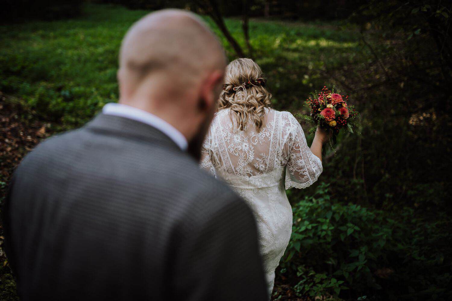 Braut im Spitzenkleid lächelt mit Blumenstrauß, Bräutigam in der Nähe – echte Hochzeitsmomente in Köln festgehalten.