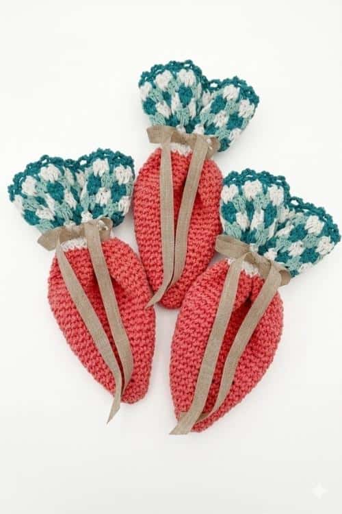 Three coral crocheted carrot pouches with green-white tops and beige ribbons are displayed on a white background. Free crochet pattern