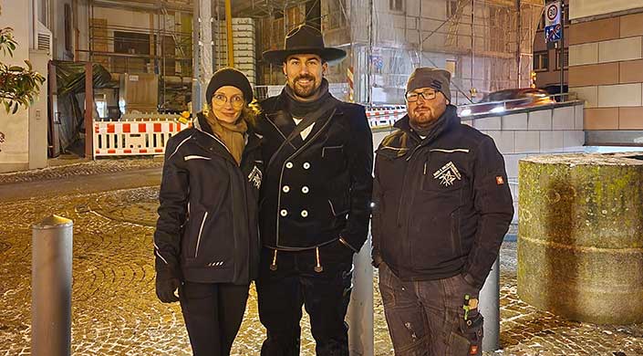 Ein Mann trägt einen traditionellen schwarzen Trachtenanzug und einen Hut, steht draußen bei Nacht und lächelt in die Kamera.