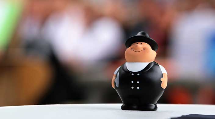 Eine kleine, rundliche Figur in schwarzer Uniform und Mütze steht auf einer weißen Oberfläche, verschwommener Hintergrund.