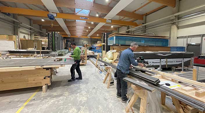 Werkstatt mit Holzbalken, Maschinen, Baumaterialien und einer Person, die arbeitet. Die Halle ist gut beleuchtet und organisiert, mit verschiedenen Werkzeugen und Arbeitsplätzen.