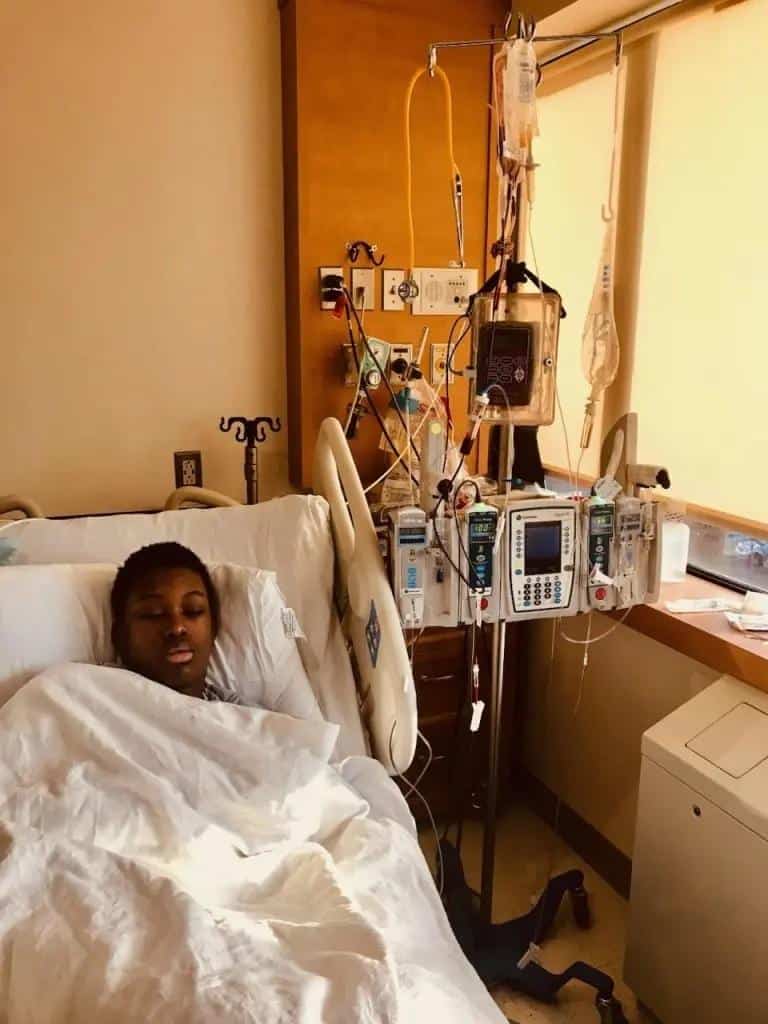 A young Black patient rests in a hospital bed, appearing at ease under a white blanket. Medical equipment, including IV drips and monitoring devices, surrounds the bed, indicating sickle cell disease treatment. This New York patient is likely receiving gene therapy for pain relief.