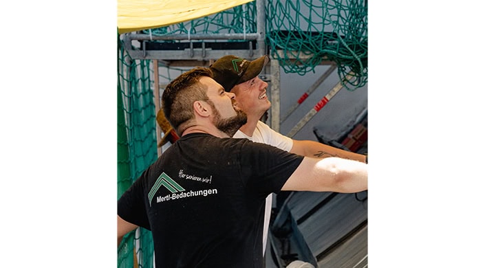 Zwei Männer arbeiten zusammen und schauen nach oben, einer trägt ein schwarzes T-Shirt, der andere ein weißes T-Shirt und eine schwarze Kappe.