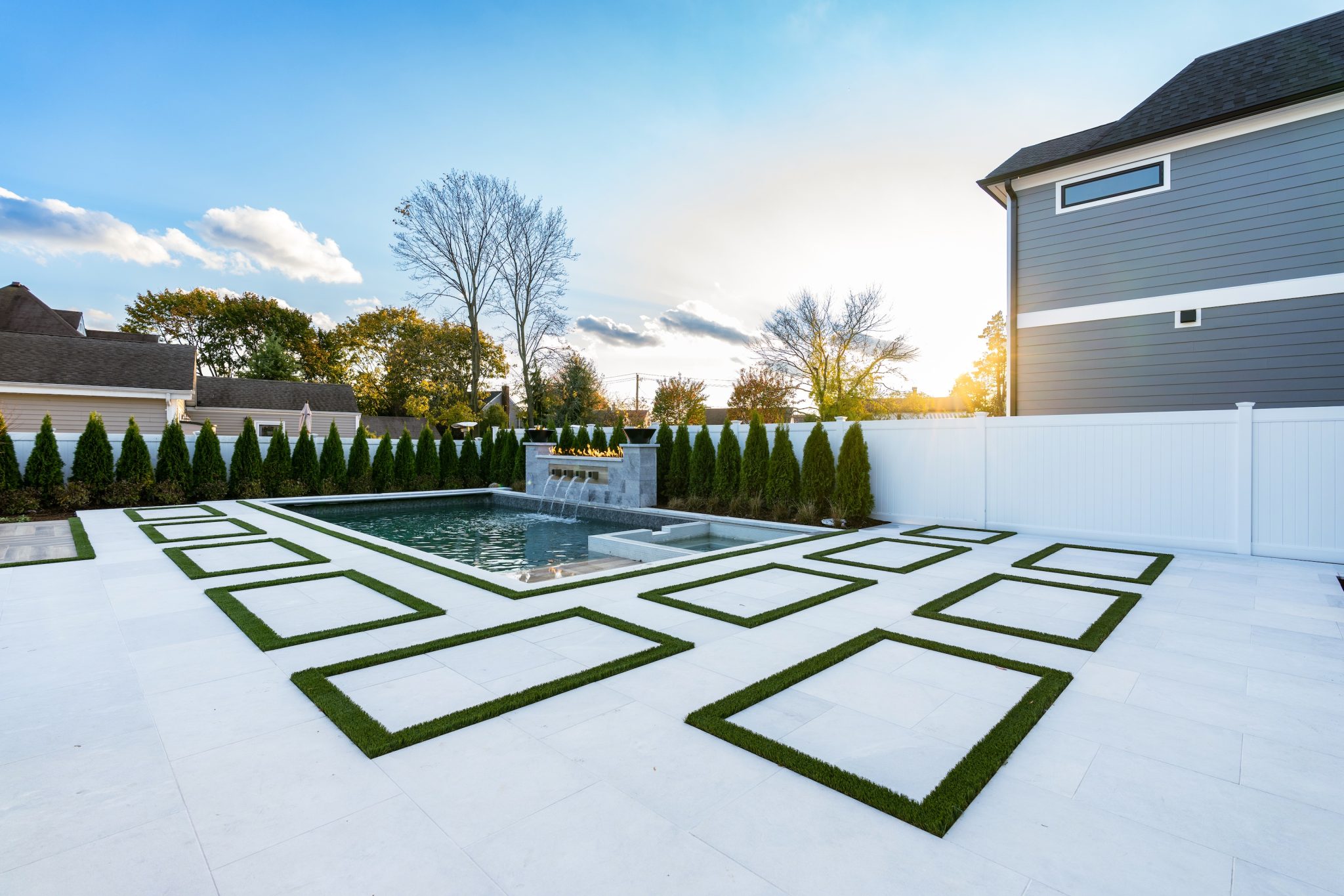 Modern backyard with a geometric patio design, rectangular pool, white fencing, and tall evergreen shrubs along the perimeter at sunset.