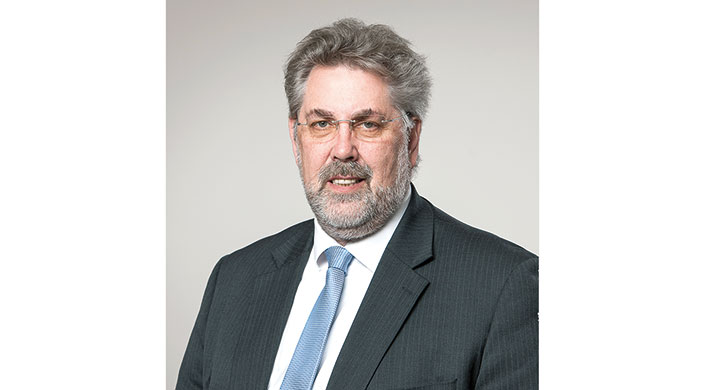 Ein älterer Mann mit grauen Haaren und Bart trägt eine Brille, einen Anzug und eine hellblaue Krawatte vor neutralem Hintergrund.