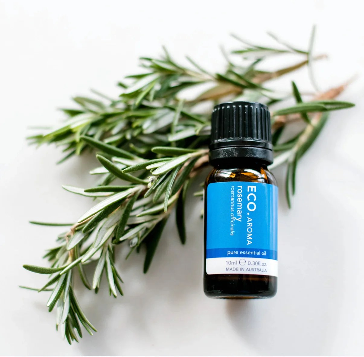 A small bottle of Rosemary Pure Essential Oil sits atop fresh rosemary sprigs on a white background, emphasizing the purity and natural essence of this product.