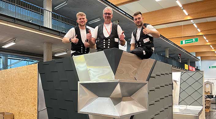 Ein Mann mit Brille und Weste steht hinter einer geometrisch geformten Metallkonstruktion und zeigt beide Daumen nach oben.