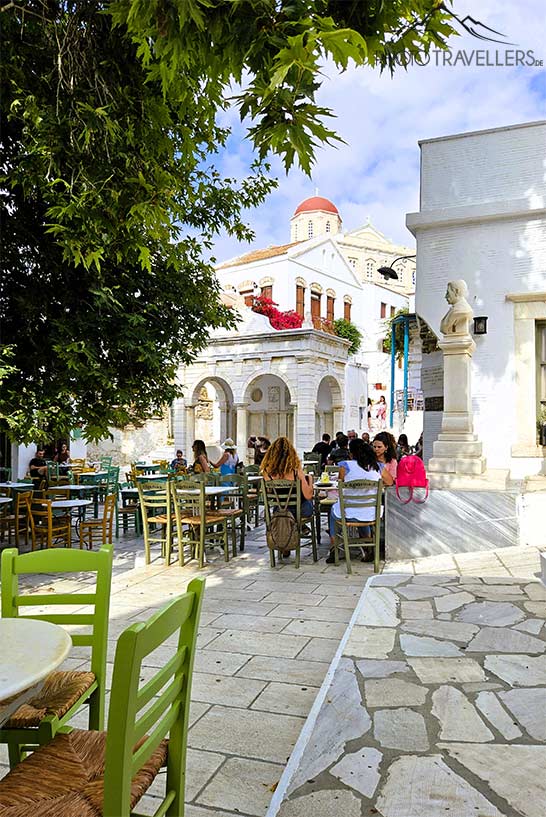 Bunte Stühle mit vielen Menschen zieren den Dorfplatz von Pyrgos rund um eine Platane