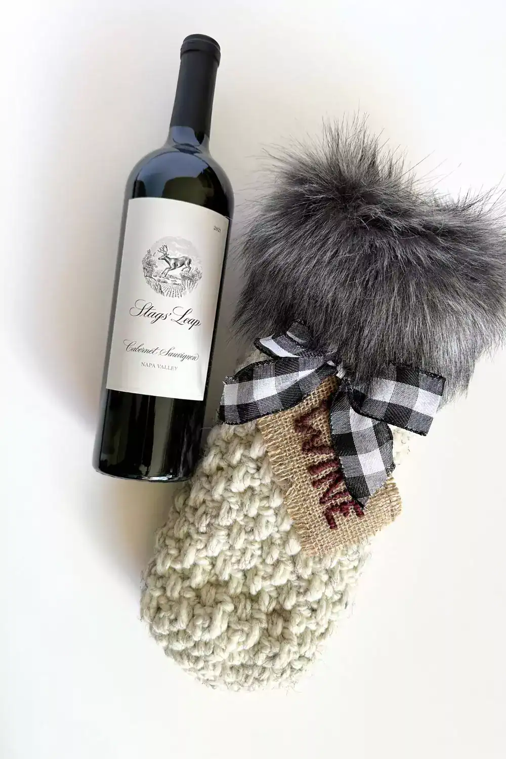 a bottle of wine and a crocheted wine bottle gift bag with faux fur fabric trim, checkered bow and wine tag