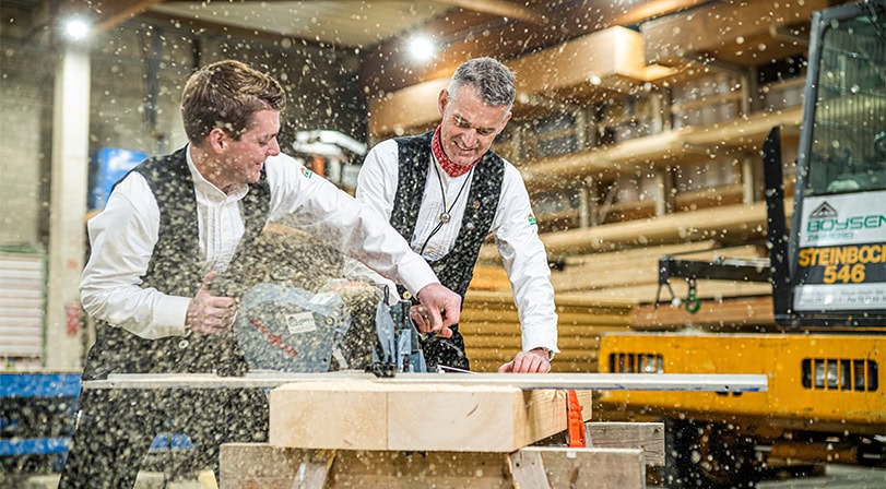 Ein Mann in traditioneller Kleidung hobelt Holz in einer Werkstatt, Späne fliegen um ihn herum.