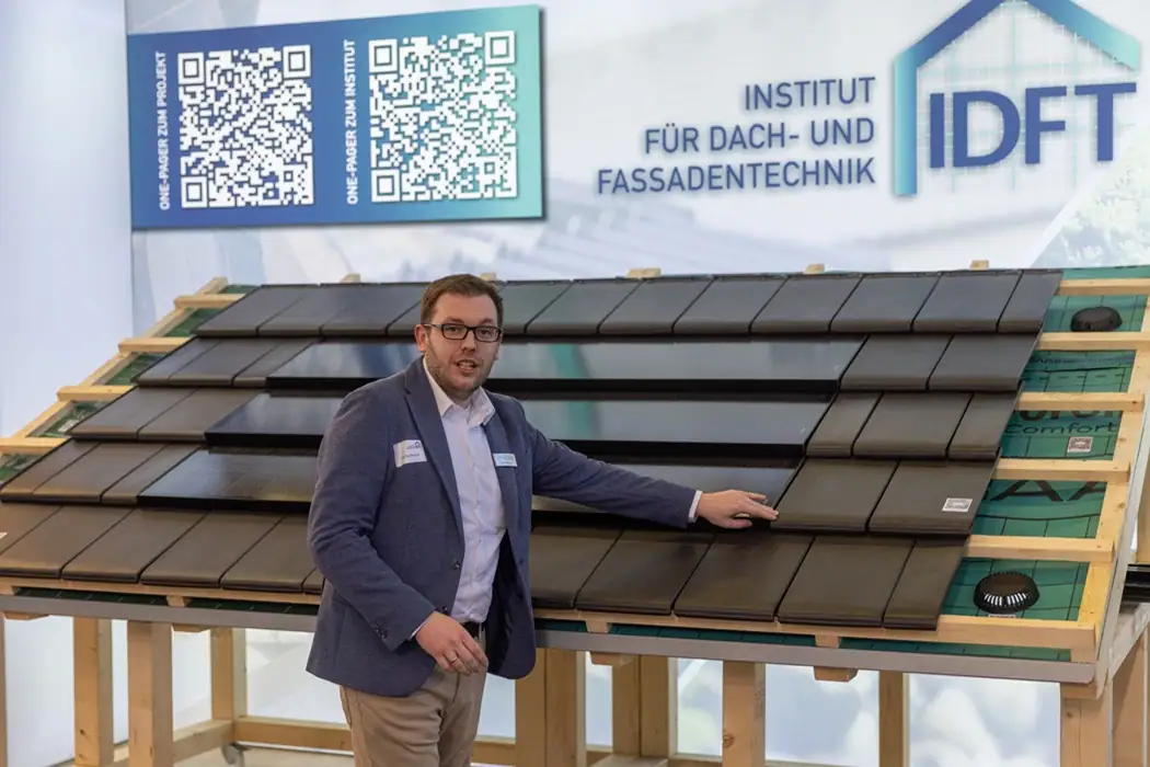Jan Redecker präsentiert ein Modell eines Dachs mit modernen Dachziegeln und PV-Indach-Modulen, im Hintergrund sind das IDFT-Logo und zwei QR-Codes sichtbar.