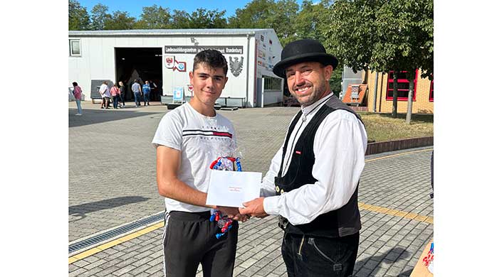 Zwei Männer stehen draußen, einer überreicht dem anderen ein Geschenk und ein Zertifikat. Sie lächeln, im Hintergrund ist ein Gebäude mit einem Wappen zu sehen.