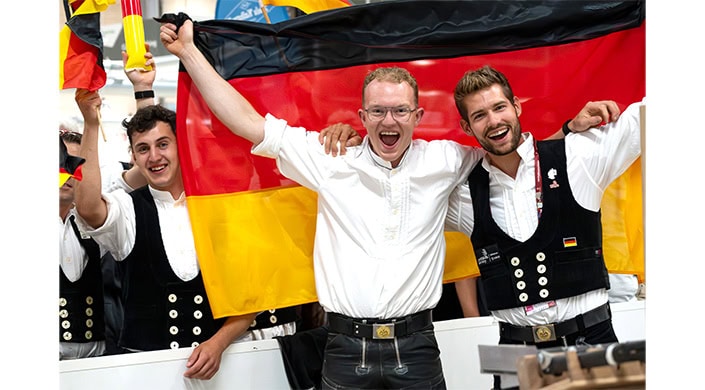 Ein lachender Mann in weißem Hemd steht vor einer großen Deutschlandflagge und hebt seine Arme, als feiere er.