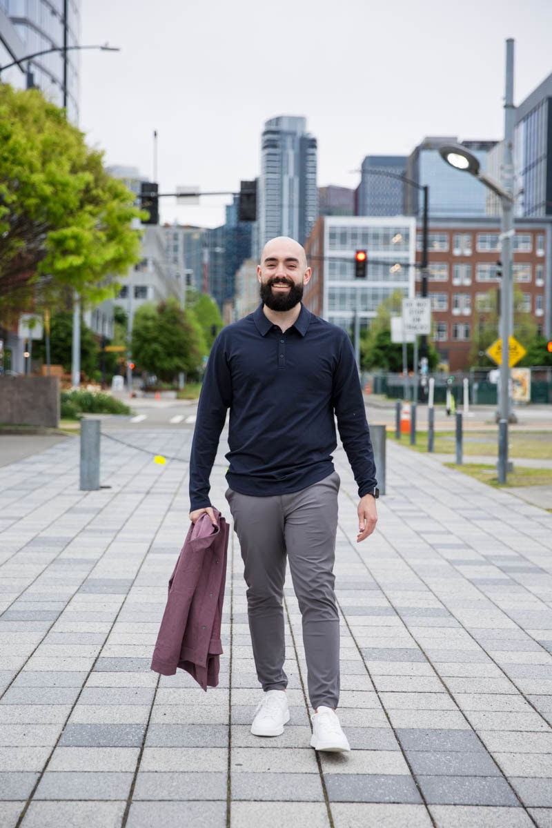 Man walking on a city sidewalk holding a jacket and smiling. Buildings and trees are visible in the background.