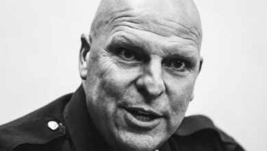 A close-up, black and white portrait of Lee County Sheriff Mike Scott, with a bald head and stern expression. He is wearing a uniform with a badge. The image captures a serious and intense moment.