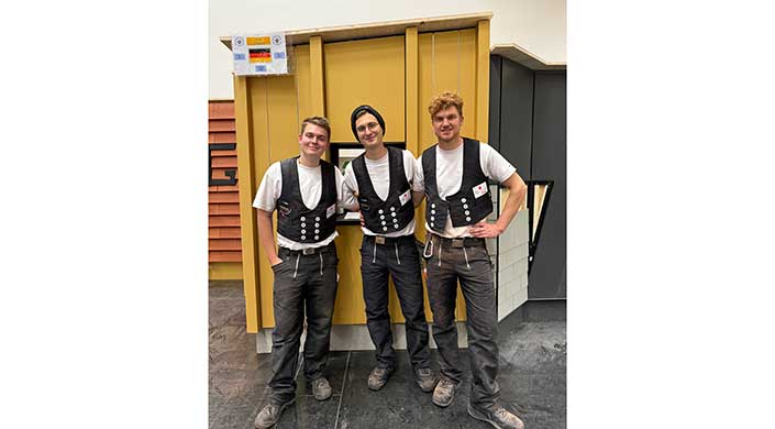 Drei junge Männer in traditioneller Handwerkerkleidung stehen lächelnd nebeneinander vor einer gelben Wand.