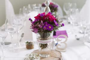 Hochzeitslocation Haus Runde 7 Eine Glasvase mit rosa und violetten Blumen schmückt einen Hochzeitstisch, neben dem Tafelaufsatz mit der Zahl 3.
