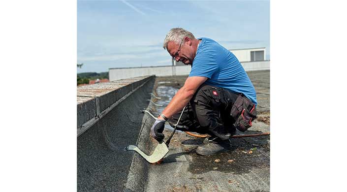 Ein Mann mit Handschuhen arbeitet an der Dachrinne eines Gebäudes und entfernt oder repariert einen Streifen Material bei sonnigem Wetter.