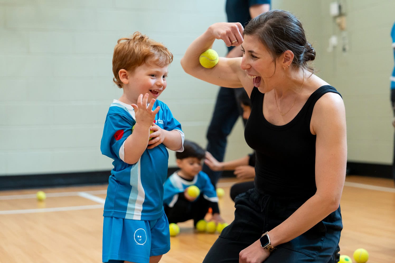 Young child in a Sportball uniform smiles with an adult during an indoor parent-participation class using tennis balls.