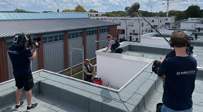 Zwei Arbeiter tragen Arbeitswesten und arbeiten auf einem Flachdach neben einem Eimer mit Werkzeug, mit Industriegebäuden im Hintergrund.