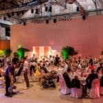 Ein Foto von edel dekorierten Tischen in der Halle. Es sitzen Hochzeitsgäste rund herum.