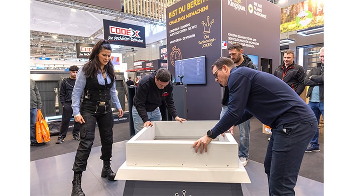 Zwei Männer bauen gemeinsam ein weißes Möbelstück auf einer Messe, im Hintergrund ein schwarzes Schild mit gelber Aufschrift.