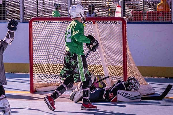 A youth hockey player in green skates past the goal as the goalie in dark gear lies on the ground inside the net.