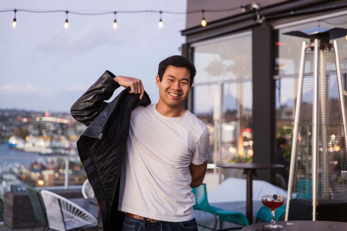 Person smiling while putting on a jacket at a rooftop bar. There are string lights above and a view of the city and water in the background.